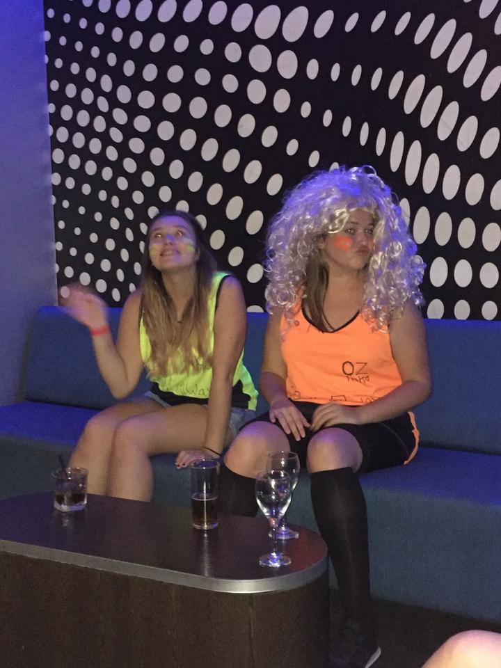 Two women in neon singlets and a curly wig pose under black-light party decor.