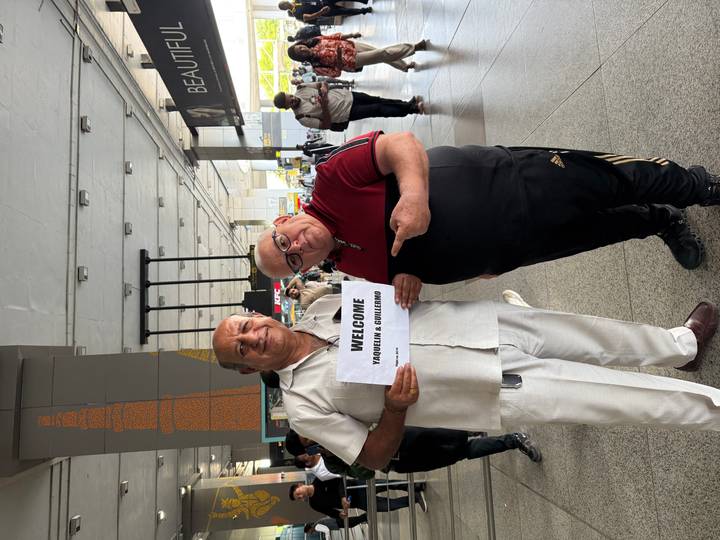 Two men posing with a welcome sign at an airport.