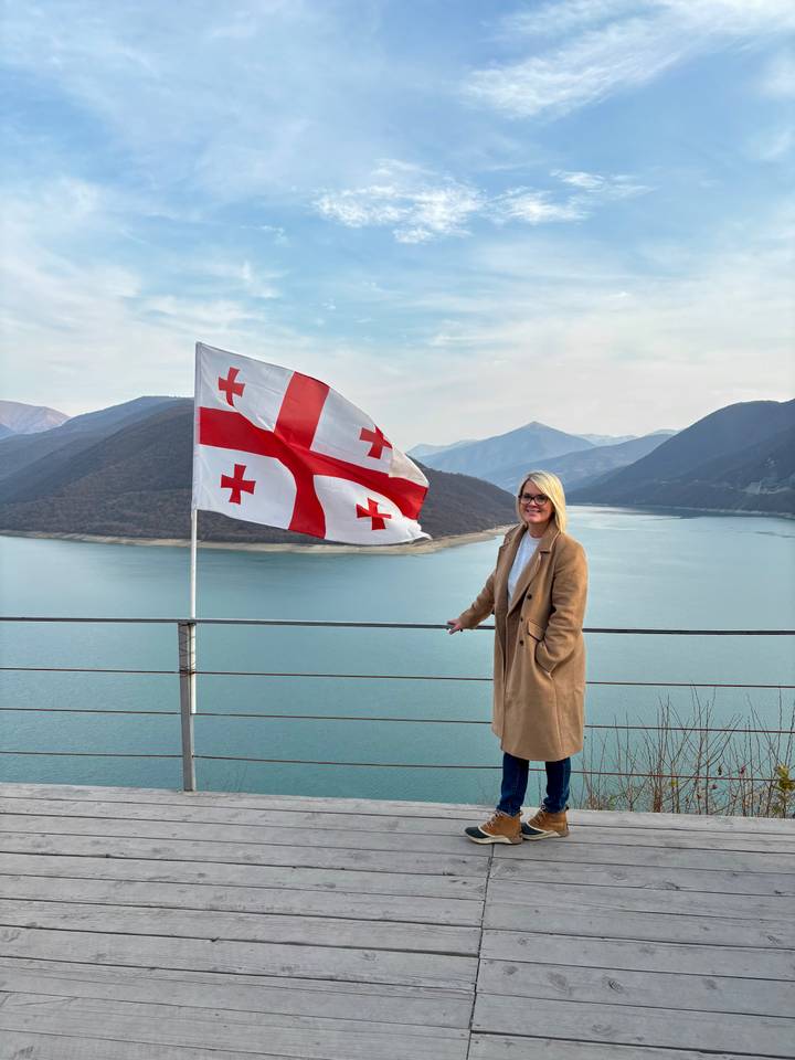 Una mujer parada junto a una bandera georgiana con montañas y un lago.