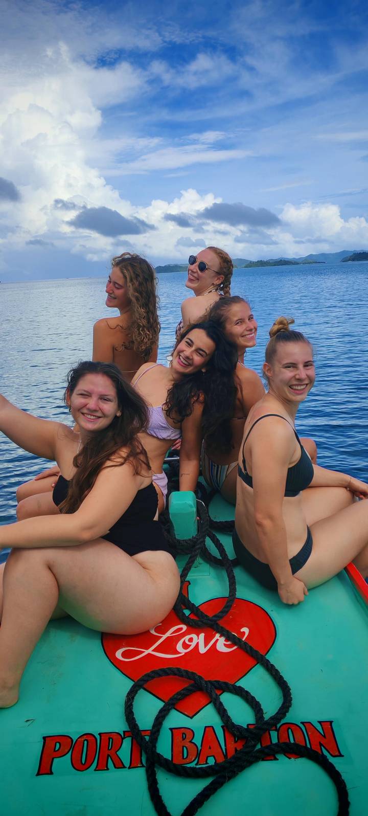 Fünf Frauen in Badeanzügen auf einem Boot in tropischer Umgebung.