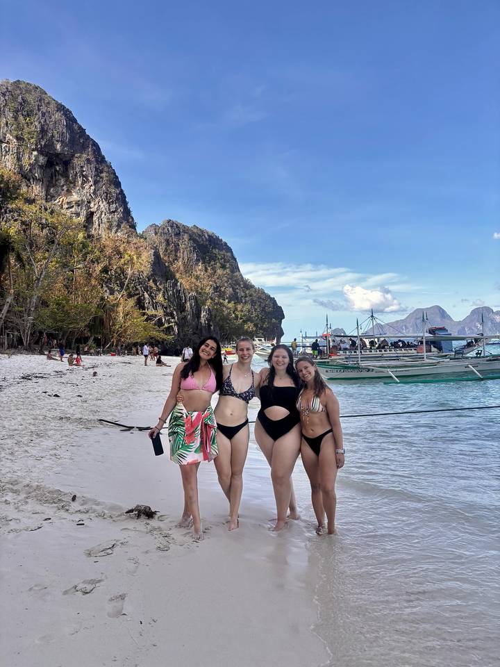 Vier Frauen posieren an einem Strand mit Klippen und Booten im Hintergrund.