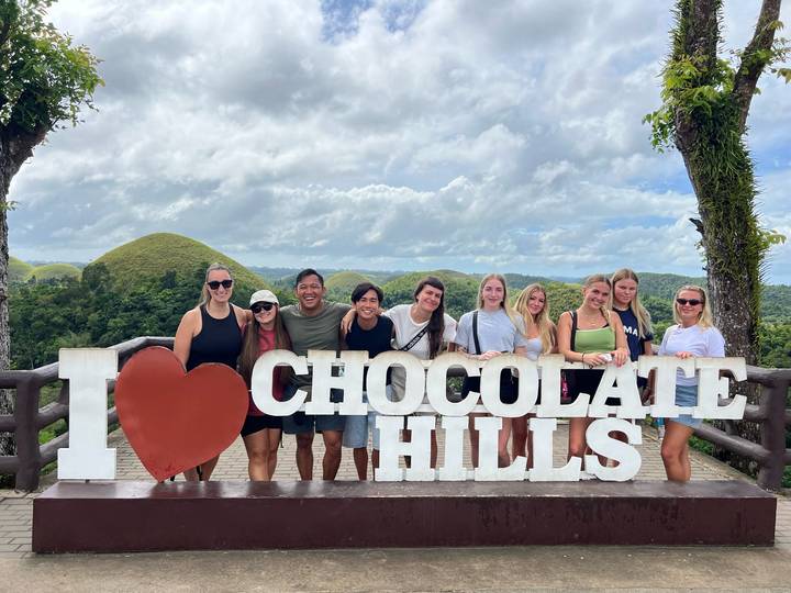 Eine Gruppe posiert mit einem "I love Chocolate Hills"-Schild mit einzigartigen Hügeln im Hintergrund.