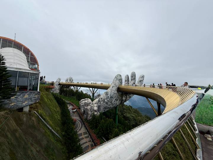 Puente dorado sostenido por manos gigantes de piedra con turistas.