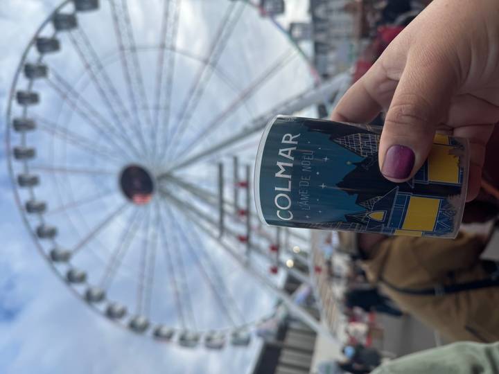 Een reuzenrad op de achtergrond met een hand die een beker vasthoudt met het label 'Colmar' op de voorgrond.
