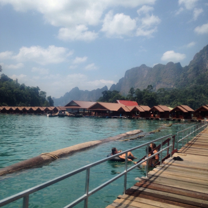 Floating wooden bungalows on turquoise lake surrounded by limestone peaks under hazy sky.
