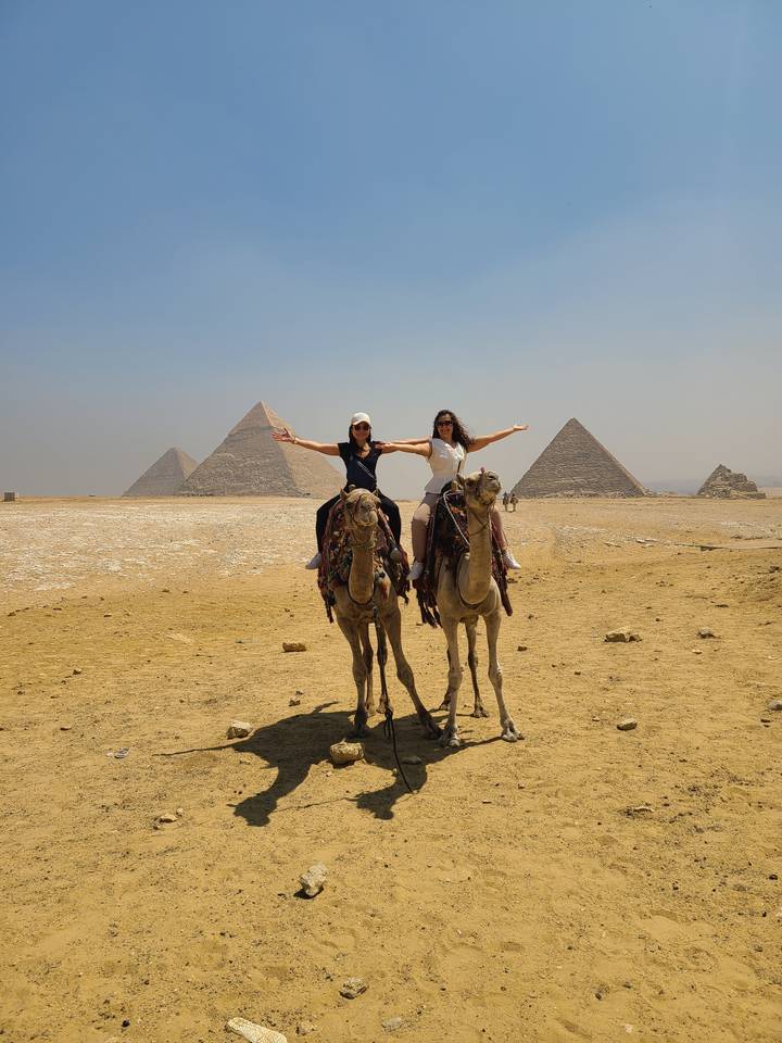 Deux personnes à dos de chameau avec des pyramides en arrière-plan.