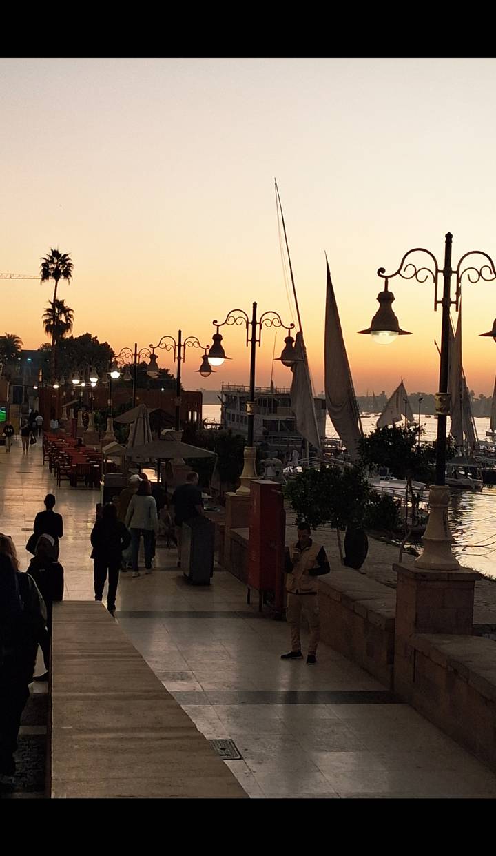 Busy riverside walkway at sunset with boats and lights.