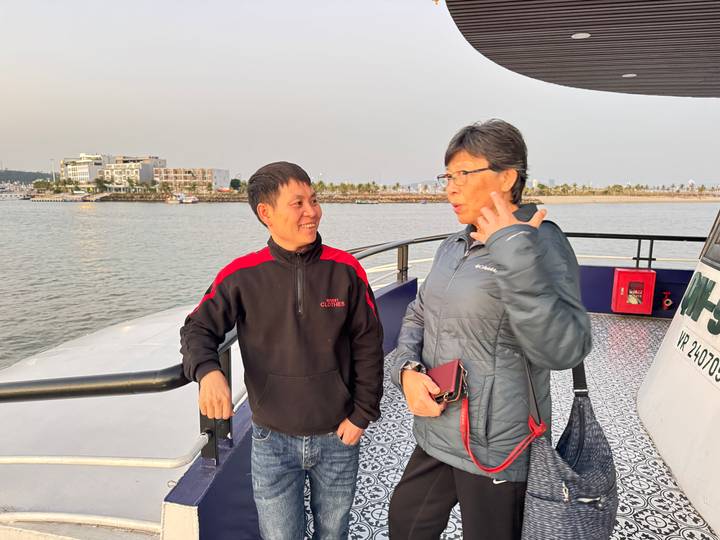 Dos personas de pie en la cubierta de un barco con agua y edificios de fondo.