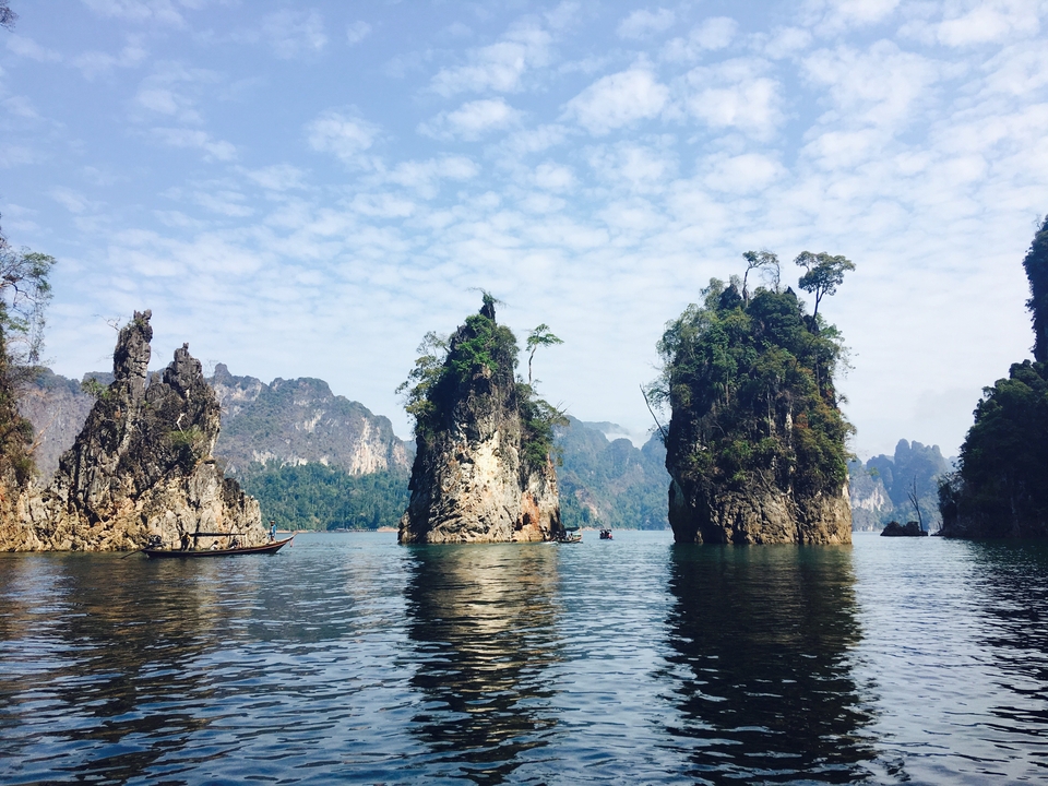 Dramatic limestone karst pillars rise from an emerald lake under a sky dotted with clouds.