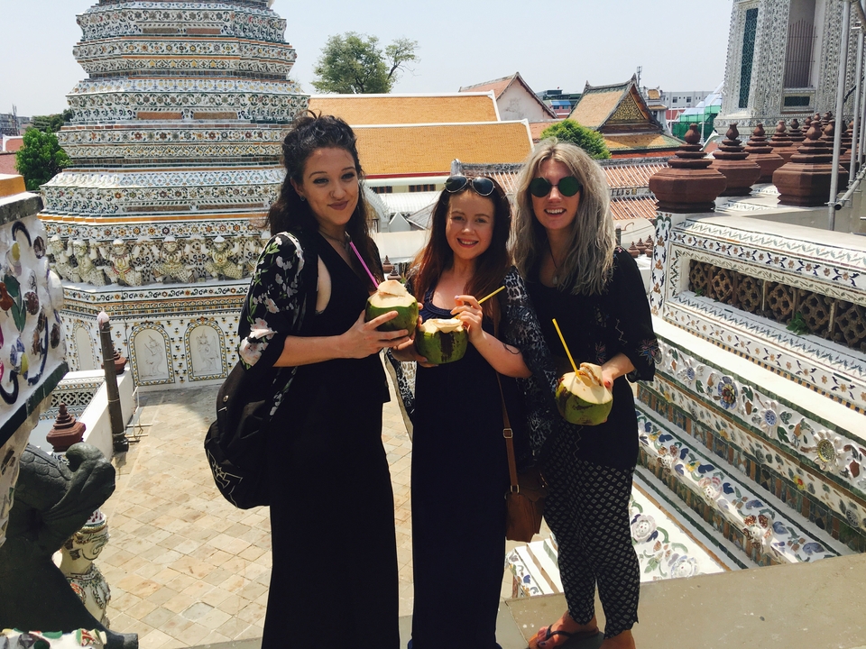 Friends holding fresh coconuts on ornate temple terrace with tiled stupas in view.