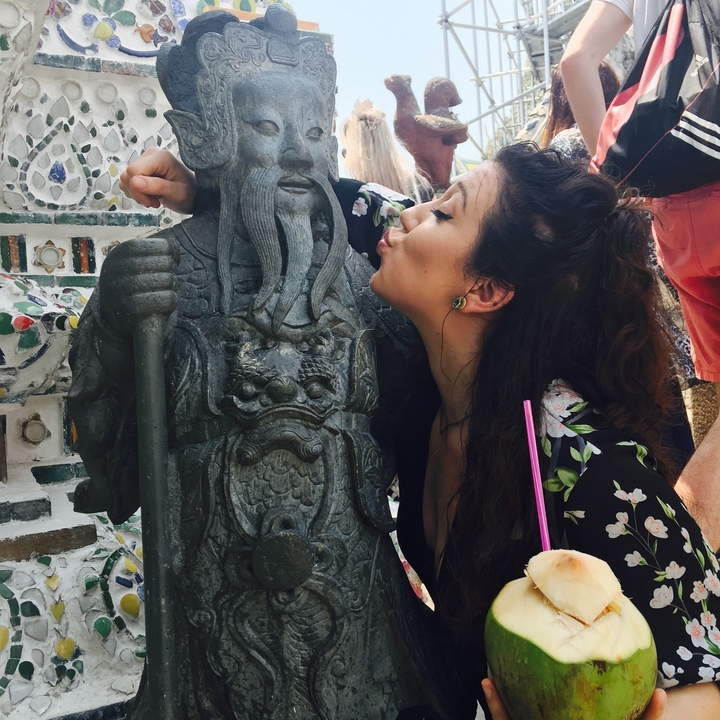 Young woman playfully kissing an ornate stone guardian statue at a Thai temple.