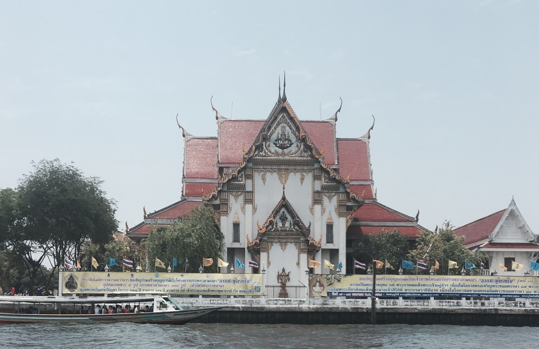 Elegant riverside temple with red roofs and ornate gables viewed across the Chao Phraya River.