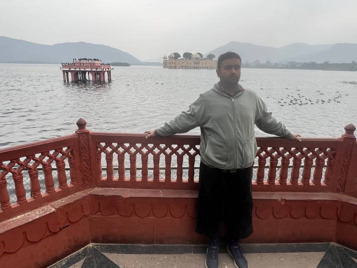 Person standing on a balcony, Jal Mahal palace and lake in the background.