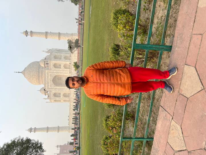 Person posing in front of the Taj Mahal with greenery in the foreground.