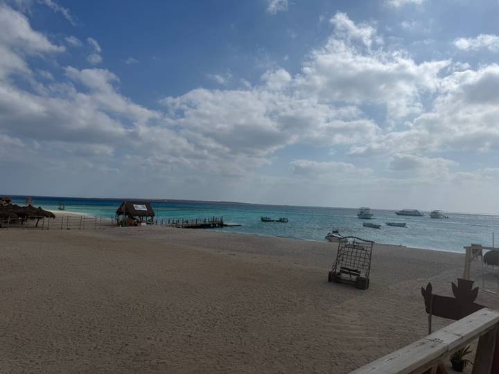 Playa arenosa con agua azul cristalina y barcos en la distancia.
