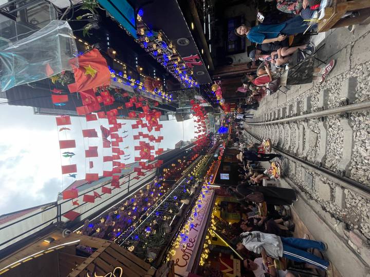 Lebendige Straßenszene mit bunten Fahnen und geschäftiger Menschenmenge entlang einer Eisenbahn.