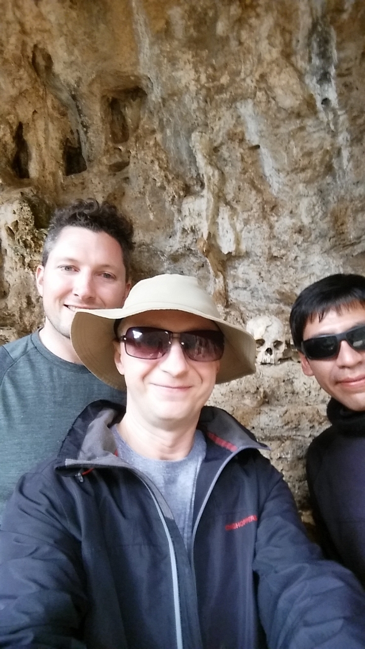 Three trekkers taking a selfie inside a cave with an ancient skull embedded in the rock behind.