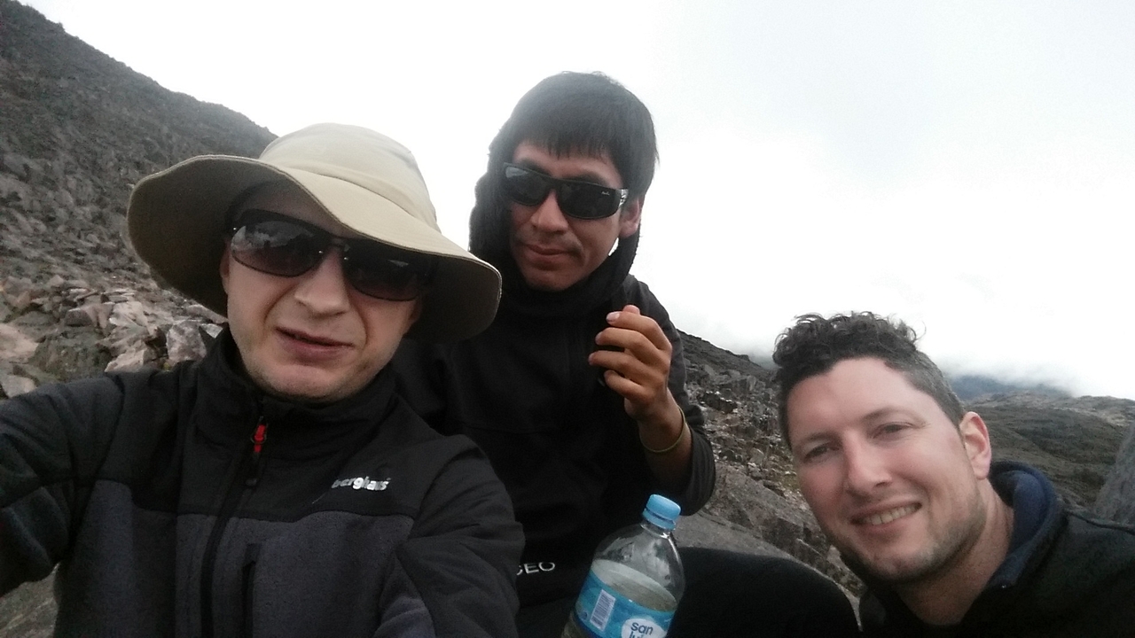 Three hikers resting on a barren rocky slope at high altitude under cloudy skies.