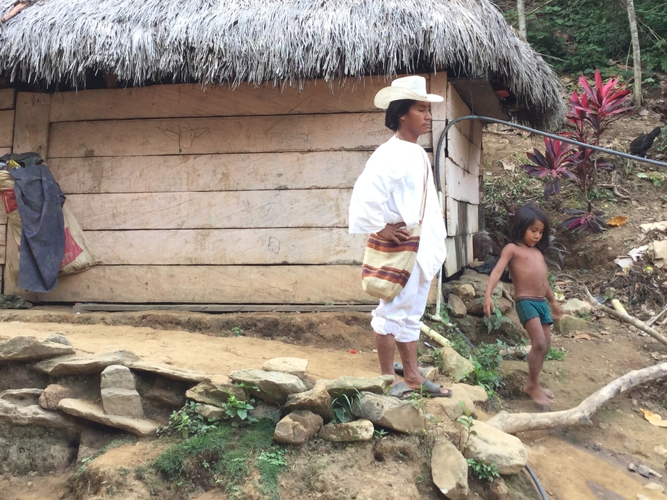 Indigenous man in traditional white attire and child standing outside a rustic wooden hut in the jungle.