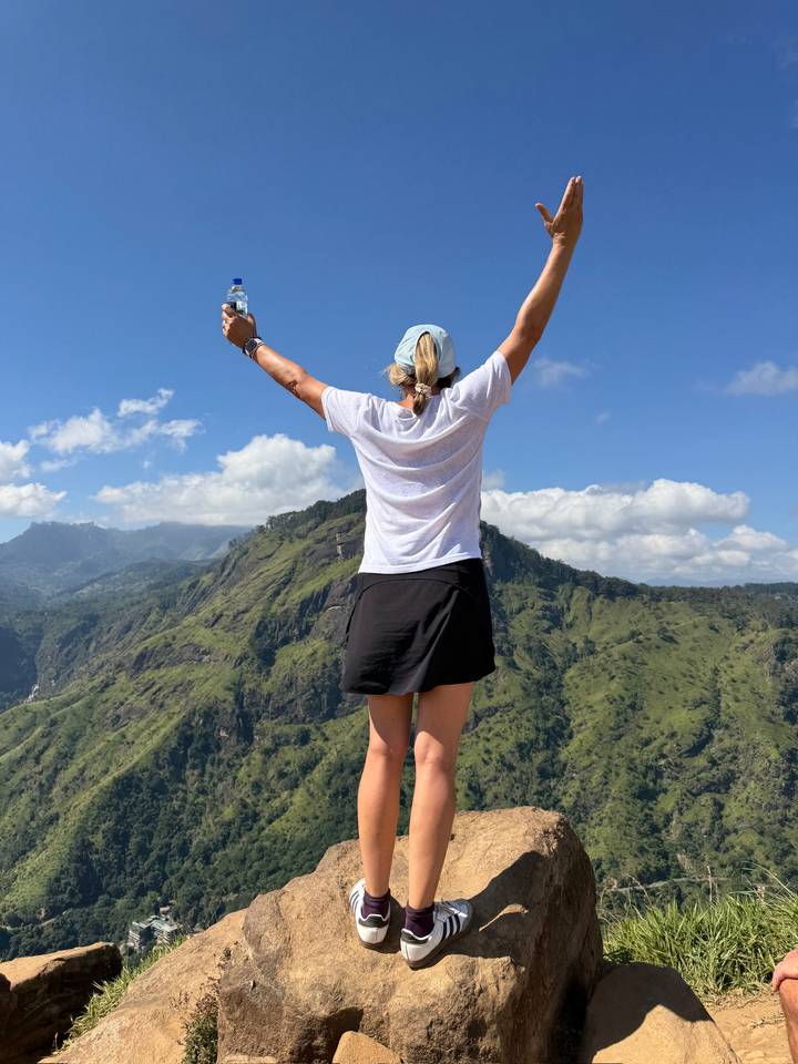 A person with outstretched arms standing on a mountain overlooking lush green hills under a bright sky.