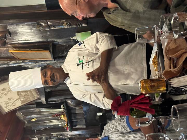 A chef in a white uniform presenting food inside a restaurant setting.