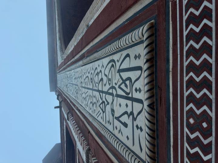 Upward perspective of intricate black marble calligraphy inlaid on a tall red-sandstone gateway panel against a clear blue sky.