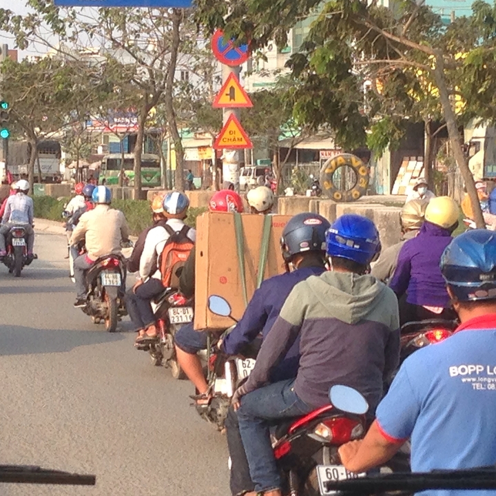 Line of motorcyclists in busy Southeast Asian traffic, some carrying boxed goods