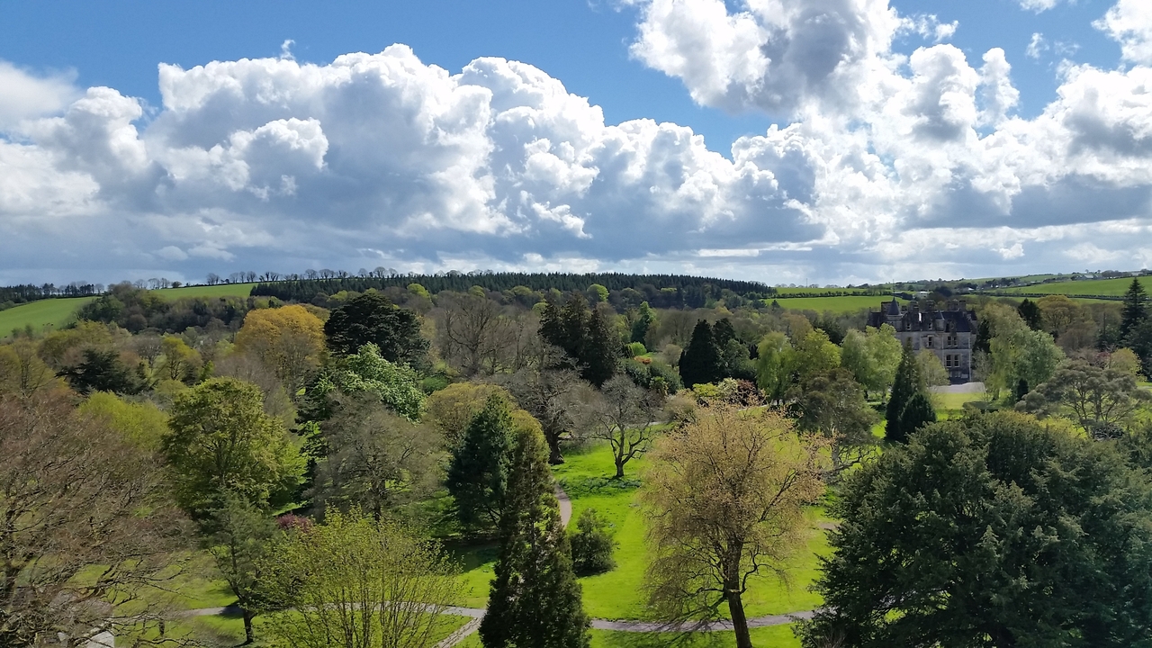 Panoramic view of a sprawling green estate with woodlands and a distant manor under billowing clouds