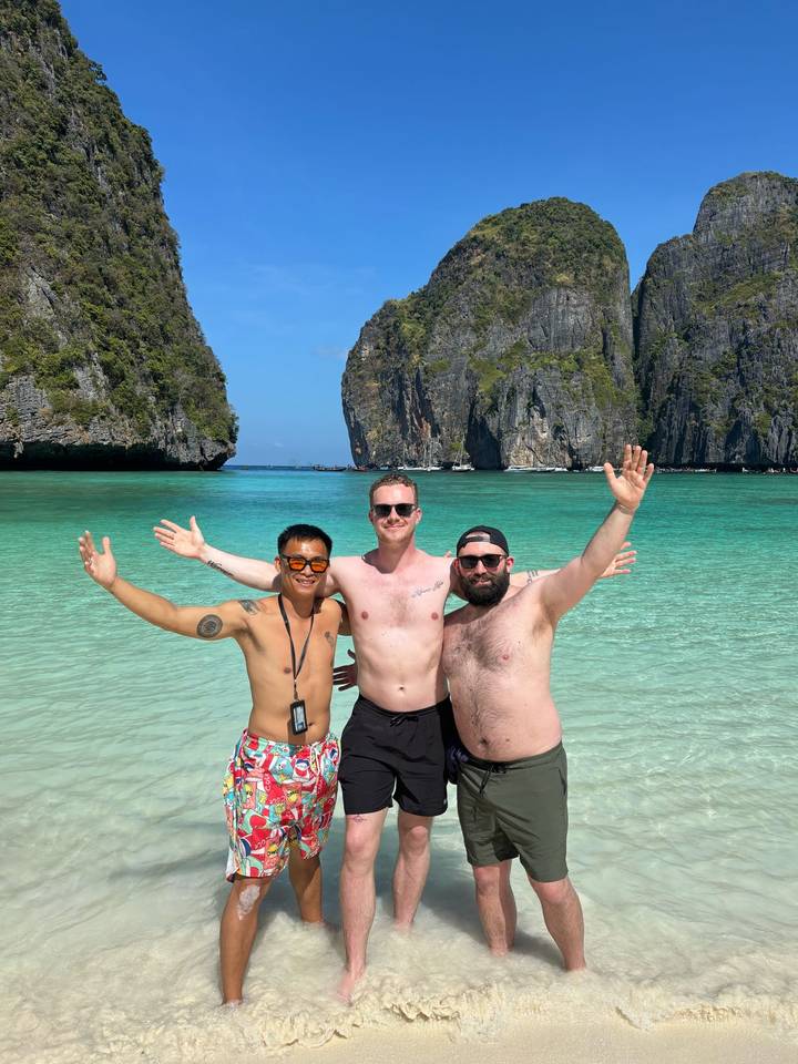Three friends with arms outstretched in crystal-clear turquoise water between dramatic limestone cliffs on a sunny day.