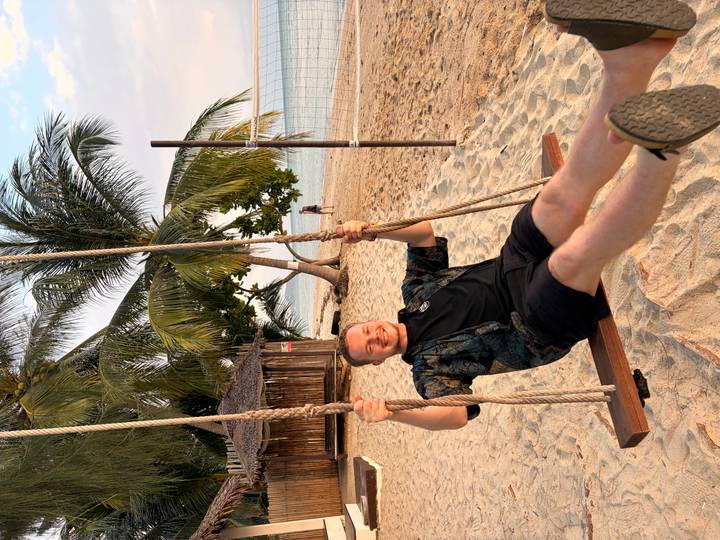 Smiling traveller swinging from a palm-tree swing on a sandy tropical beach at sunset.