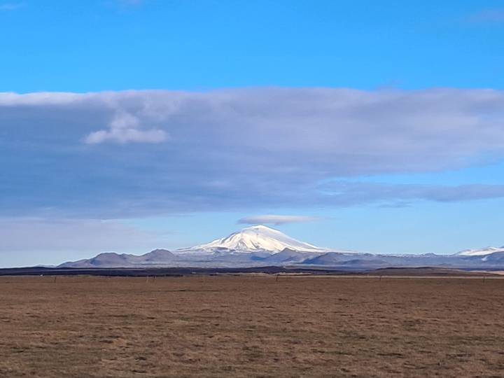Snow-covered stratovolcano rises above barren brown plains beneath a broad cloud bank.