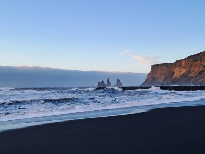 Waves crash on black sand beach with dramatic Reynisdrangar sea stacks and cliff at dusk.