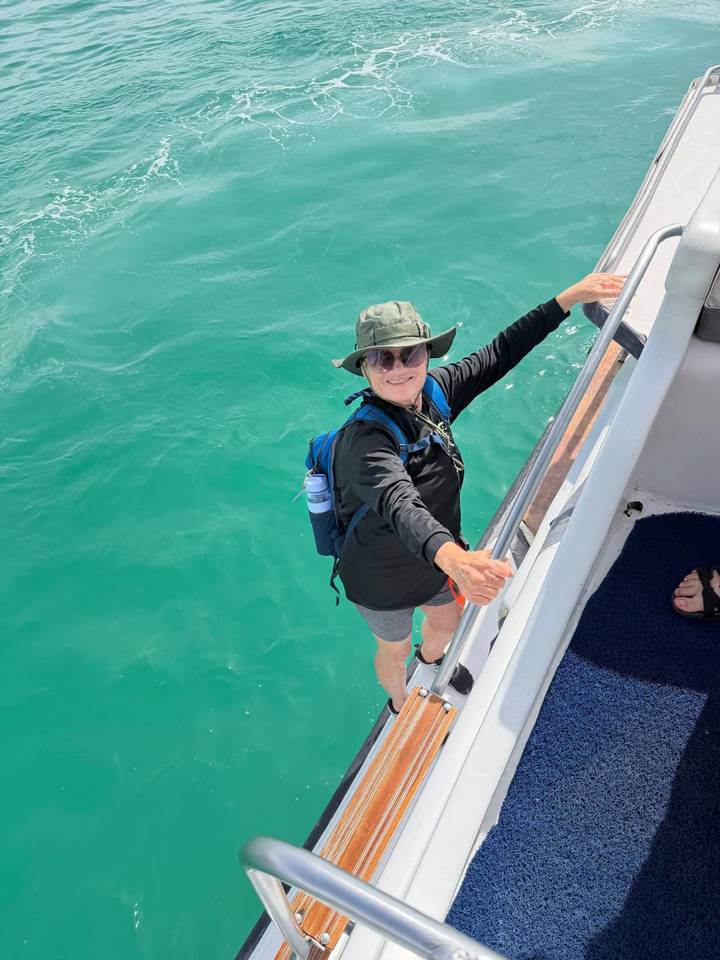 Woman in hat stepping from a boat ladder into clear turquoise water, wearing a backpack.