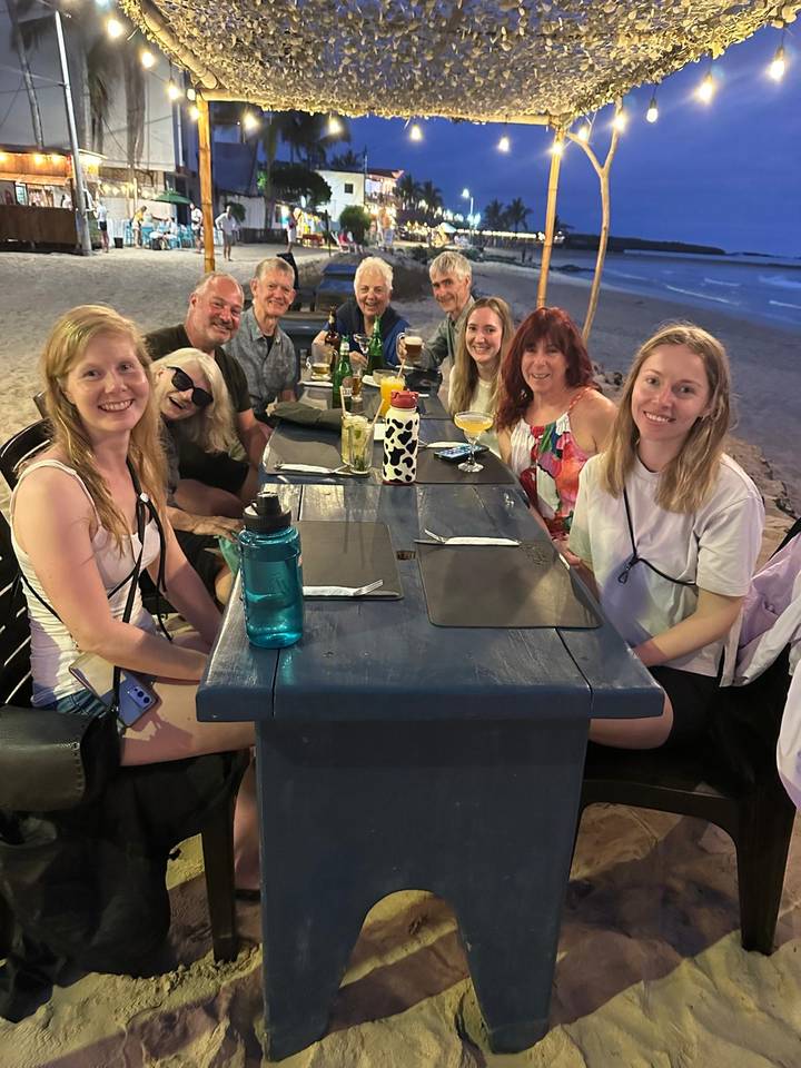 Travel group sharing drinks around a blue wooden table at an open-air beach restaurant at night.