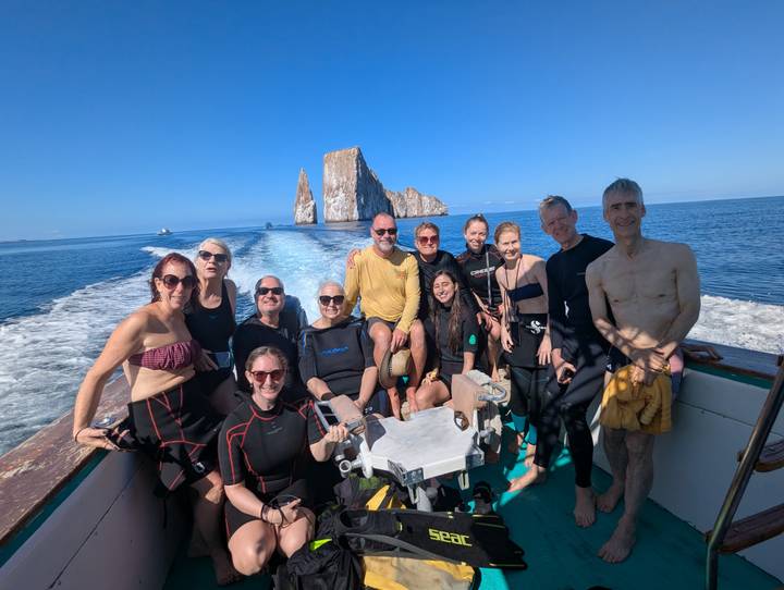 Group in wetsuits posing on a boat with the dramatic Kicker Rock rising behind in open sea.