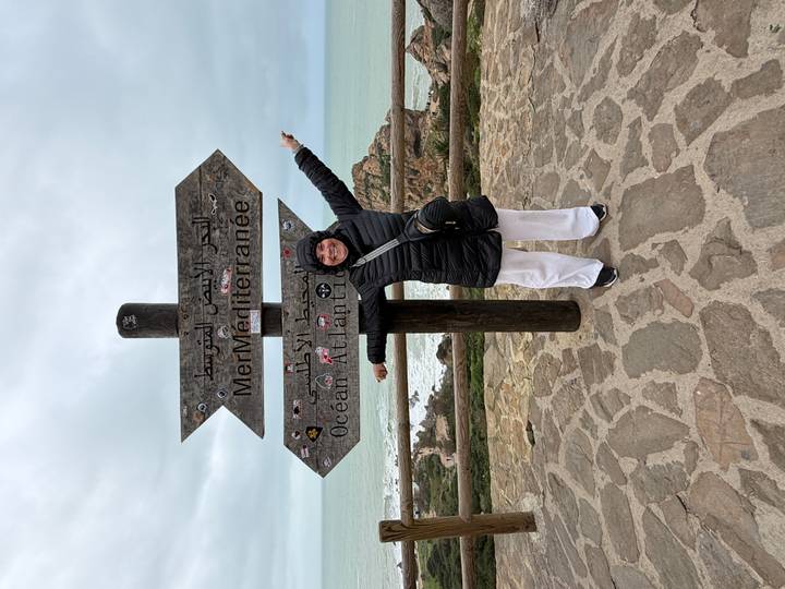 Traveller points happily at a bilingual sign marking the Atlantic and Mediterranean seas with cliff and ocean beyond.