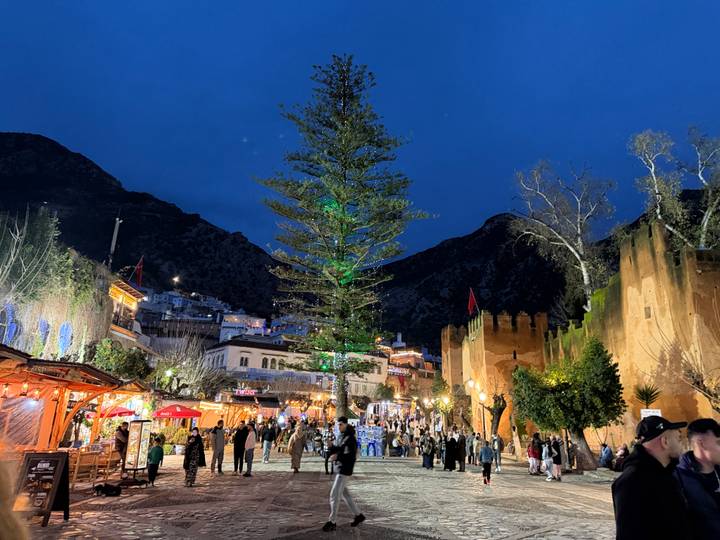 Evening market square alive with people, lights and a tall lit tree against mountain backdrop.