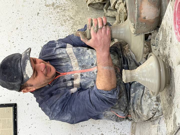Artisan potter shapes a clay tagine on a wheel, covered in grey splatters of wet clay.