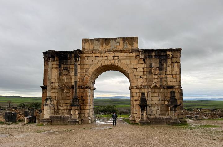Ancient Roman triumphal arch stands alone on a green plain under grey skies with a lone visitor below.
