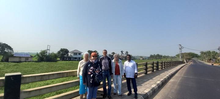 Five travelers pose on a rural bridge with open fields and village houses behind them under a bright sky.