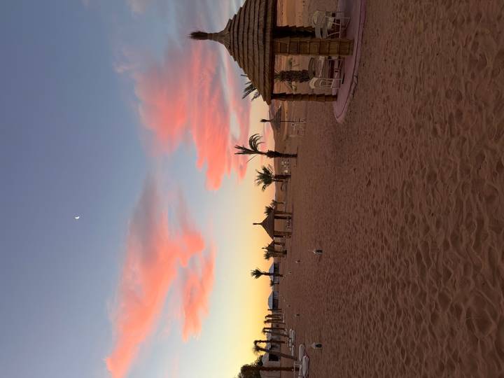 Wide sandy desert camp at dusk with scattered palms, lanterns and pink clouds overhead.