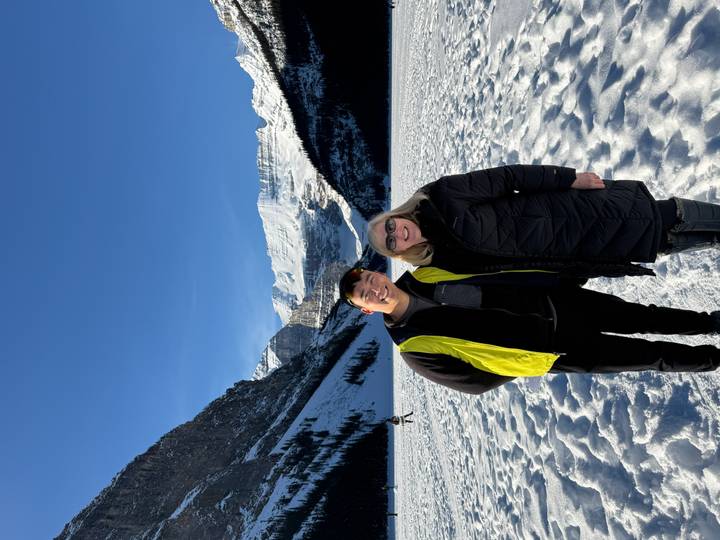 Two smiling tourists pose on the snowy surface of a frozen alpine lake with towering peaks behind.