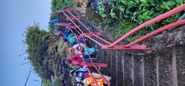 Pilgrims in colorful clothing ascend a steep concrete staircase bordered by red railings and greenery.