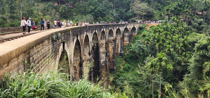 Tourists walk along the famous Nine Arches railway bridge spanning a lush green valley.