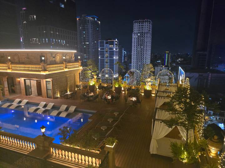 Stylish rooftop restaurant and infinity pool at night overlooking a modern city skyline with guests dining under string lights.