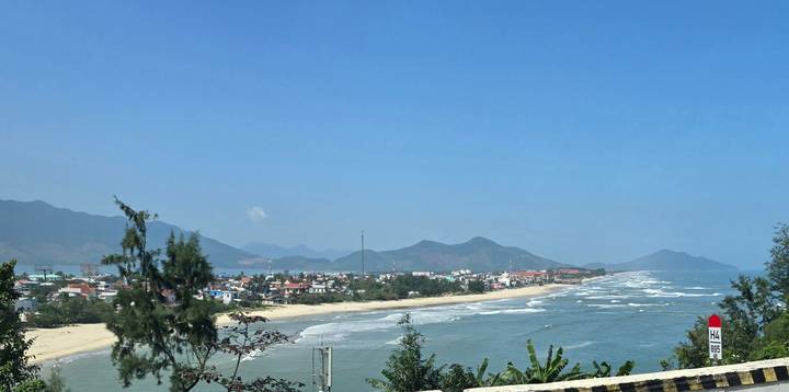 Panoramic coastal view of a long sandy beach, waves, and a small seaside town backed by hills under a clear blue sky.