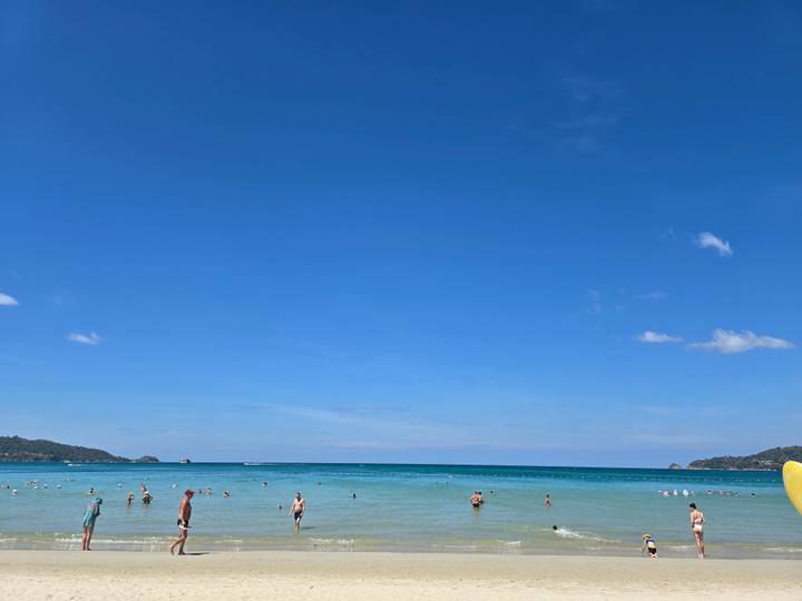 Wide sunny beach with turquoise water and scattered swimmers beneath a vast clear blue sky.