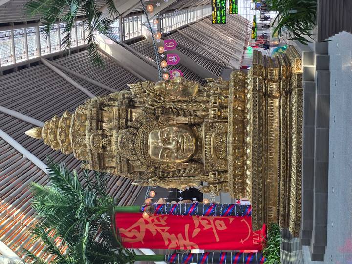 Large golden replica of Bayon faces displayed inside a modern terminal with high wooden ceiling beams.