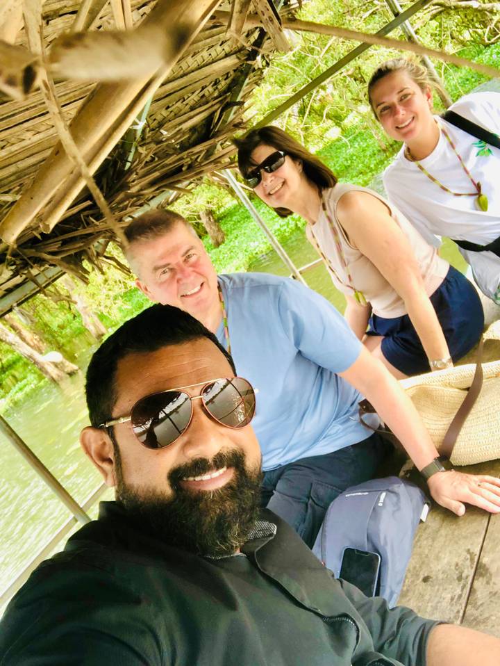 Selfie of three people on a small covered boat cruising through green waters with lush riverbank vegetation.