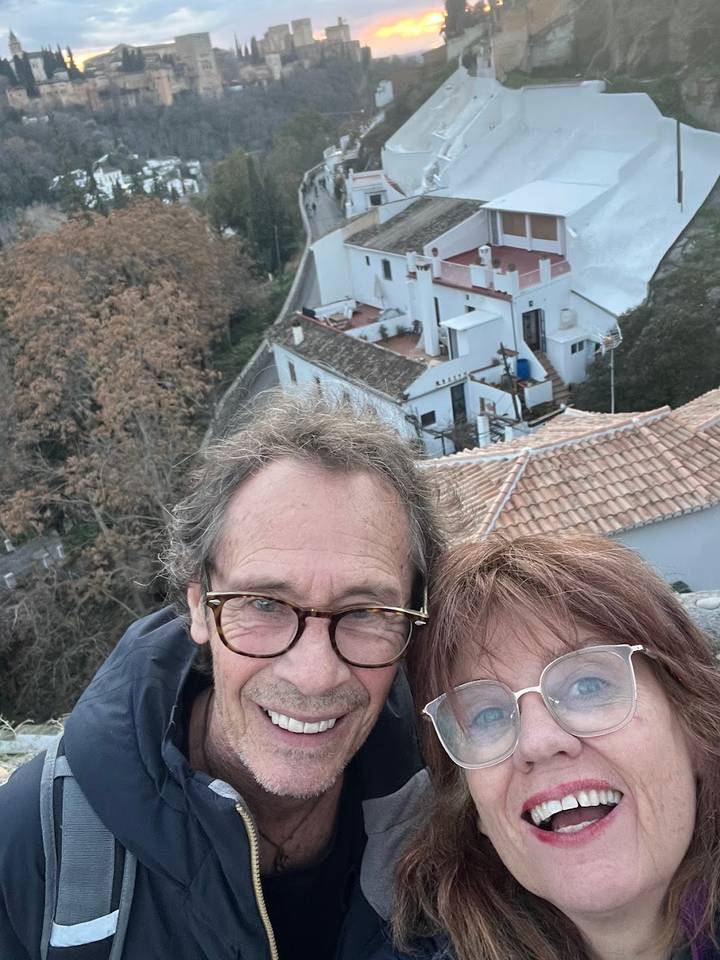 Close selfie of a smiling couple with tiled rooftops and a winding hill road in the background.